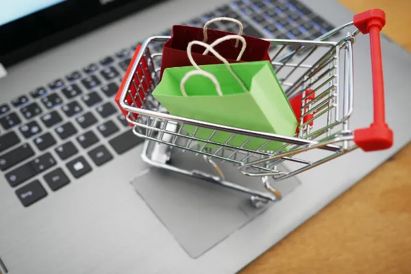 A miniature silver shopping cart containing a small red and green shopping bag, sitting on a silver laptop keyboard.