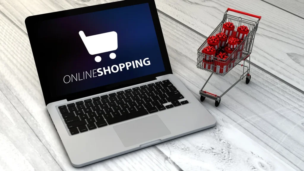 A silver laptop displaying an "Online Shopping" logo on the screen sits next to a miniature metal shopping cart filled with small red gift boxes on a light wood surface.