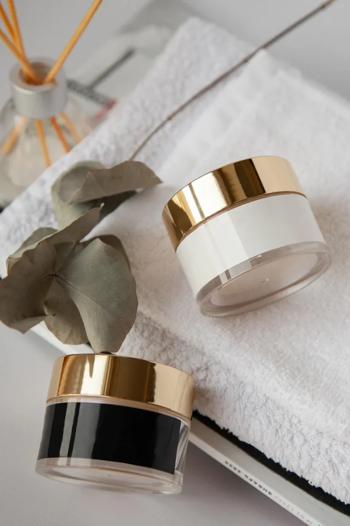 Two minimalist cosmetic jars with gold lids resting on a folded white towel next to a eucalyptus branch and a reed diffuser.