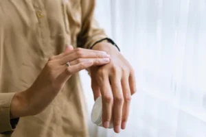 A close-up shot of a person’s hands applying a white cream or moisturizer from a small jar onto the back of their hand near a bright window.